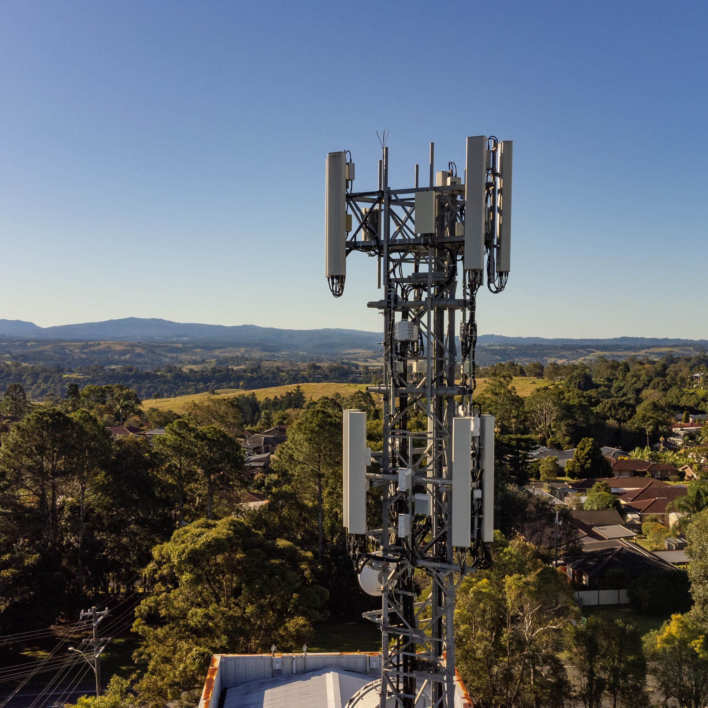 Drone-captured image of a telecommunications tower during aerial inspection service, with surrounding suburban landscape and hills in the background.